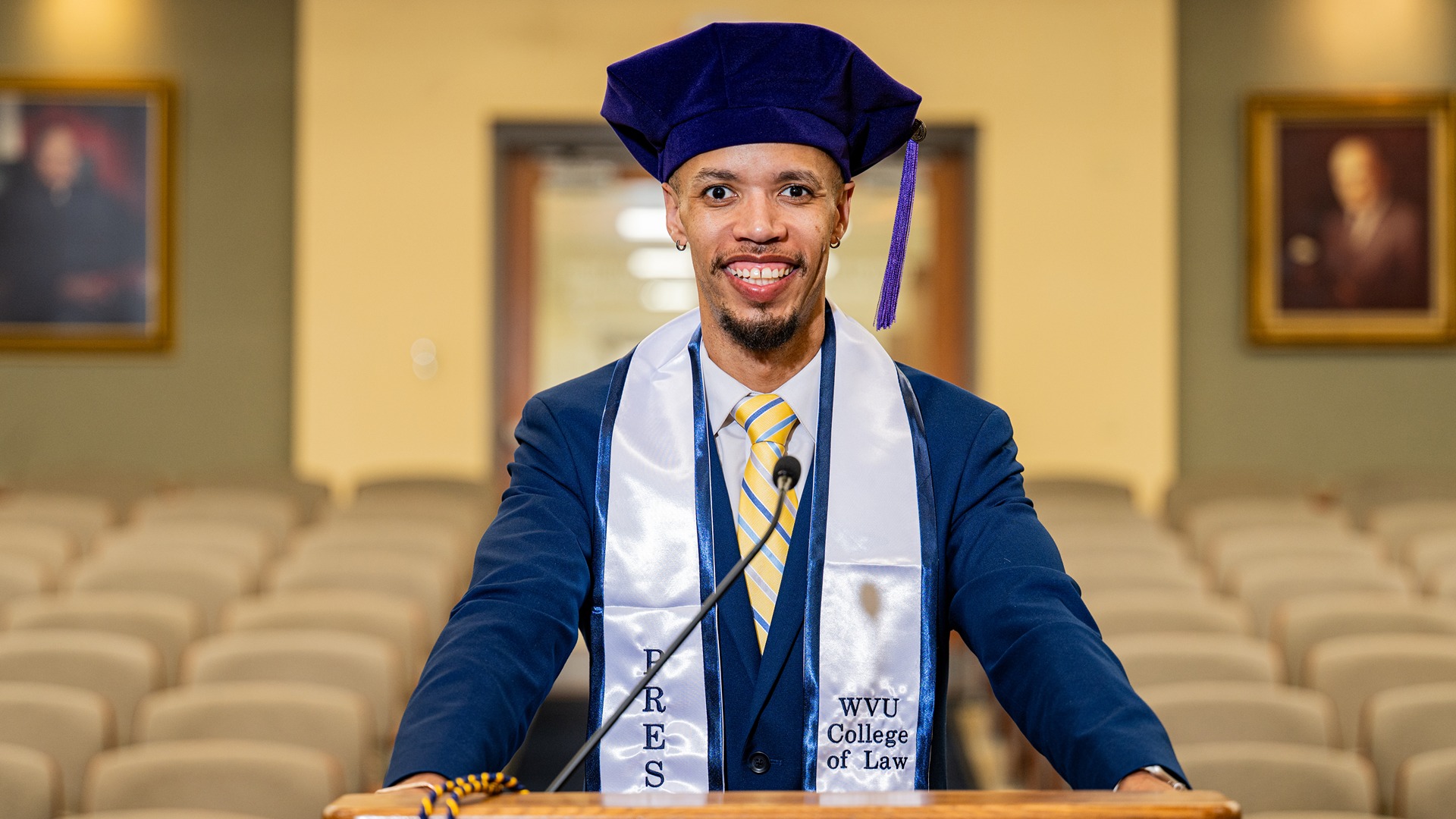 Photo of Chris Payne in graduation regalia posing at a podium.