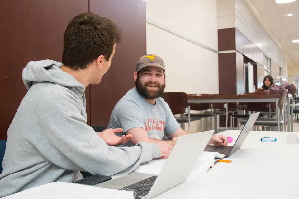Two students sit at a table at the College of Law conversing while typing on their laptops.